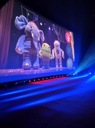 Uma tela de Cinema com luzes coloridas na base. No canto direito da tela estão projetados três logos de conferences. No centro da tela há um elefante azul, um Android e um robô com símbolo da Apple. Possivelmente mascotes das tecnologias envolvidas no evento.